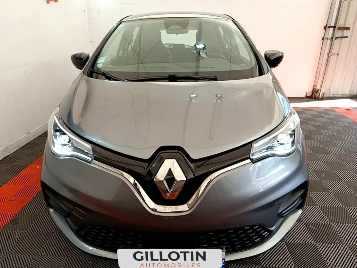 Vue frontale d'une Renault ZOE gris clair avec phares LED allumés dans un garage fermé.