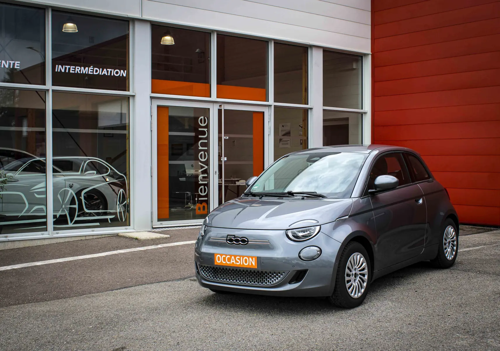 Fiat 500 électrique gris minéral métal vue 3/4 avant avec plaque orange "OCCASION" devant un bâtiment moderne.