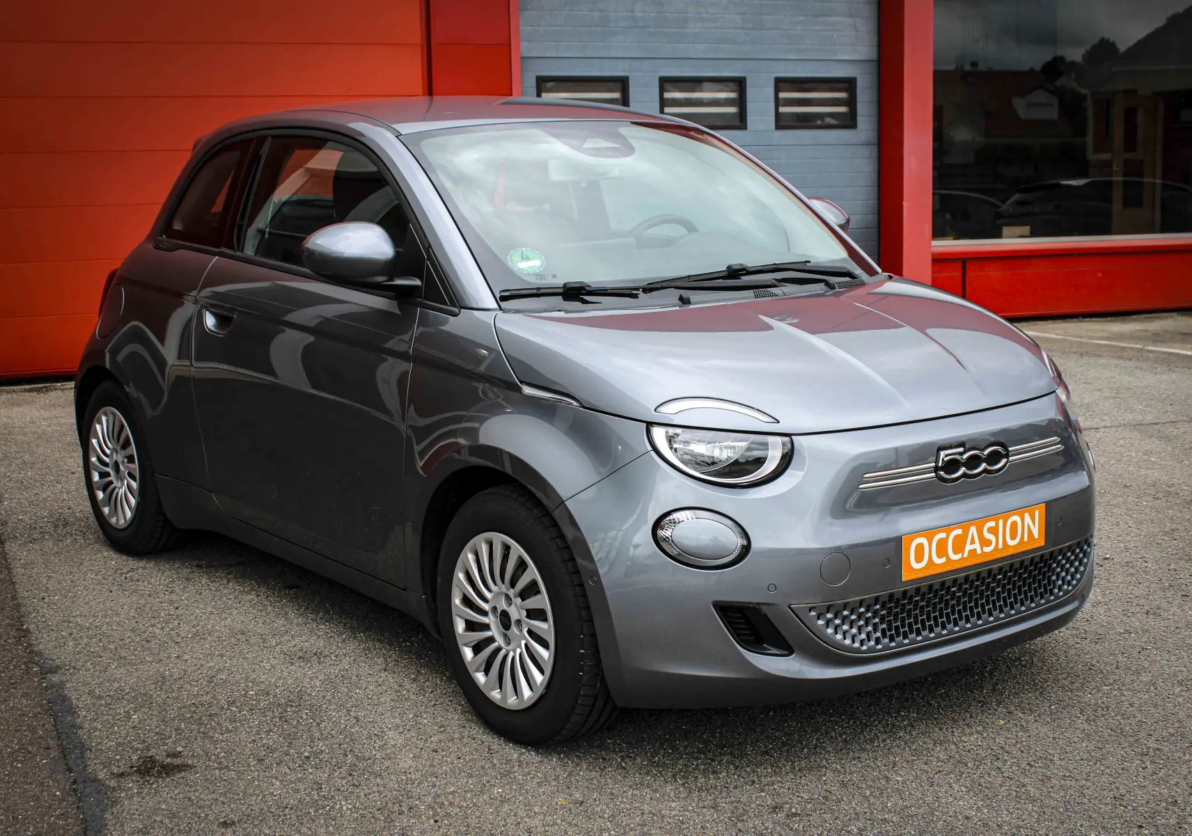 Fiat 500 électrique gris minéral métal vue 3/4 avant droit avec plaque orange "OCCASION" devant un mur rouge.