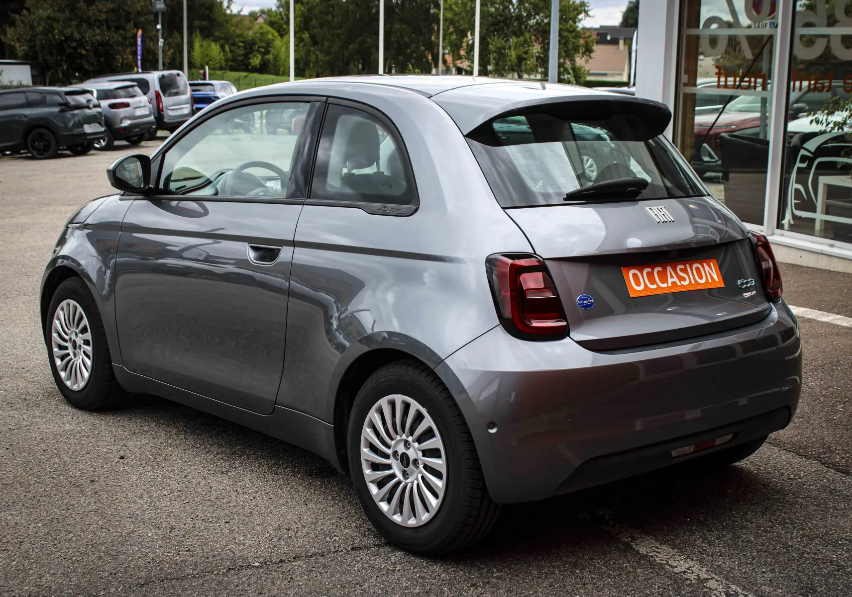 Vue 3/4 arrière droite d'une Fiat 500 électrique gris minéral métal avec jantes à rayons et badge "Occasion" sur le coffre.