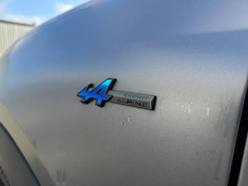 Gros plan sur l'emblème bleu et noir "Esprit Alpine" sur la carrosserie gris schiste d'un Renault Scénic électrique.