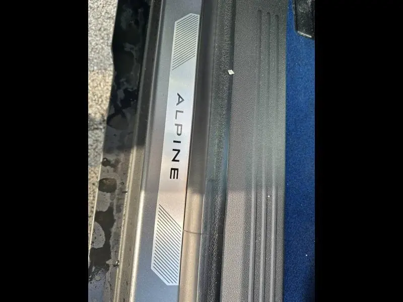 Gros plan sur la plaque de seuil de porte avec inscription Alpine, intérieur avec moquette bleue visible.