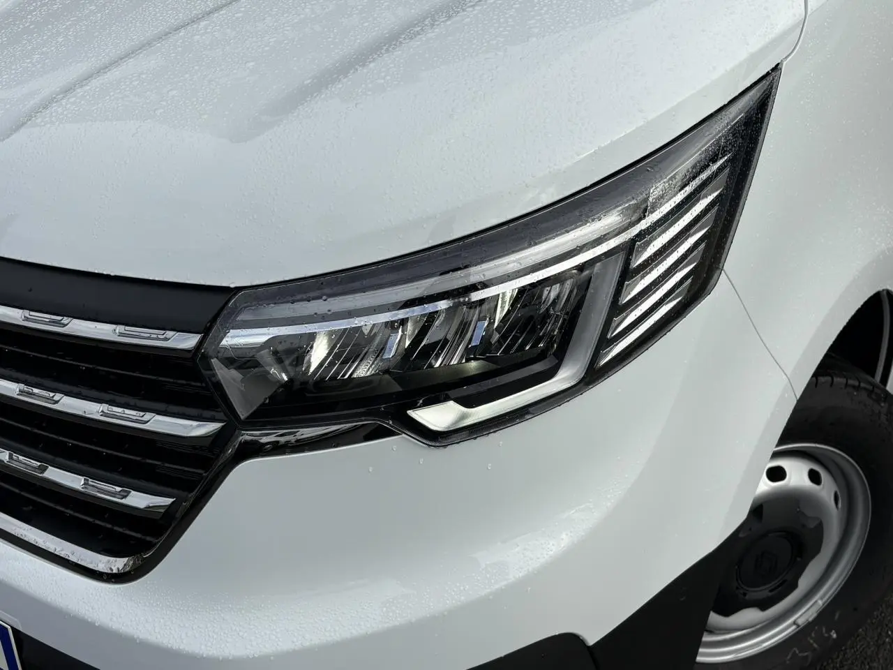 Gros plan sur le phare avant droit et la calandre chromée du Renault Trafic Fourgon blanc glacier, avec gouttes de pluie visibles.