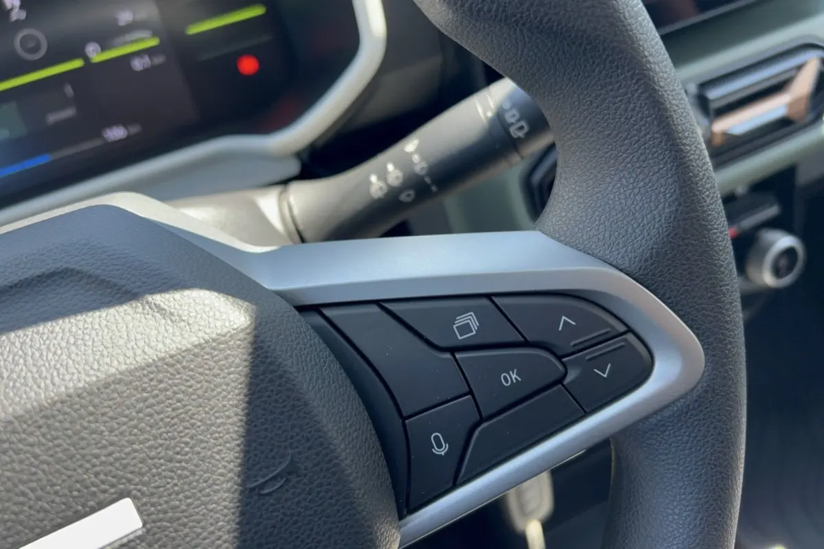 Gros plan sur les commandes au volant côté droit du Dacia Spring gris éclair, avec tableau de bord numérique en arrière-plan.