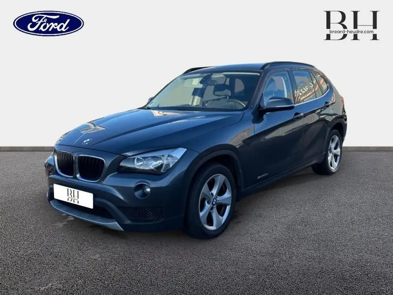 BMW X1 sDrive20d 2013 Mineralgrau vue 3/4 avant avec barres de toit noir mat et jantes alu 17 pouces.