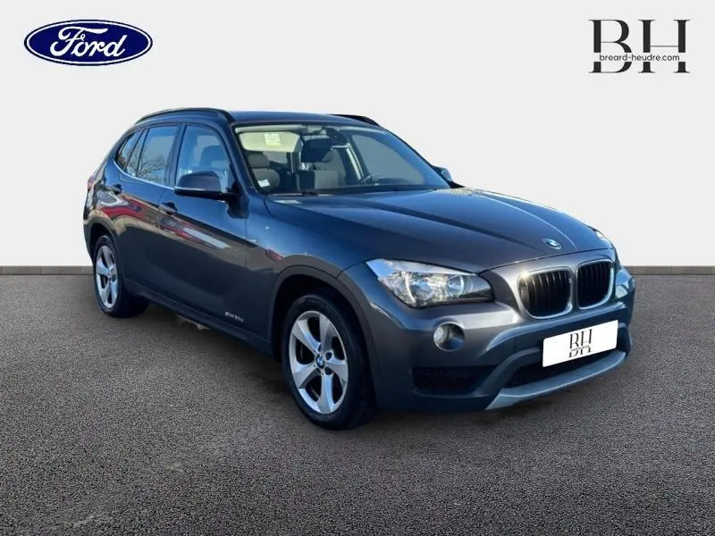 BMW X1 sDrive20d gris Mineralgrau vue 3/4 avant droit avec barres de toit noires et jantes alu 17 pouces.
