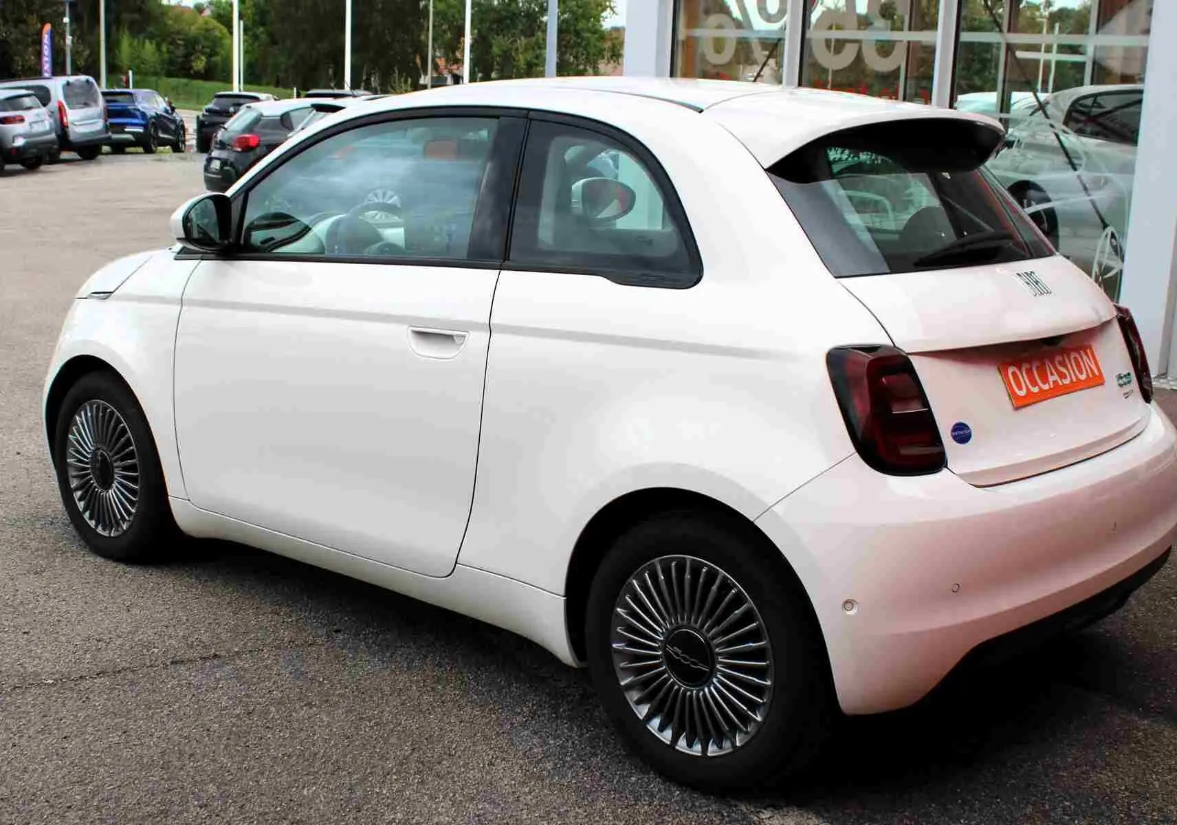 Vue 3/4 arrière droite d'une Fiat 500 électrique blanche avec jantes spécifiques et plaque "OCCASION" visible.
