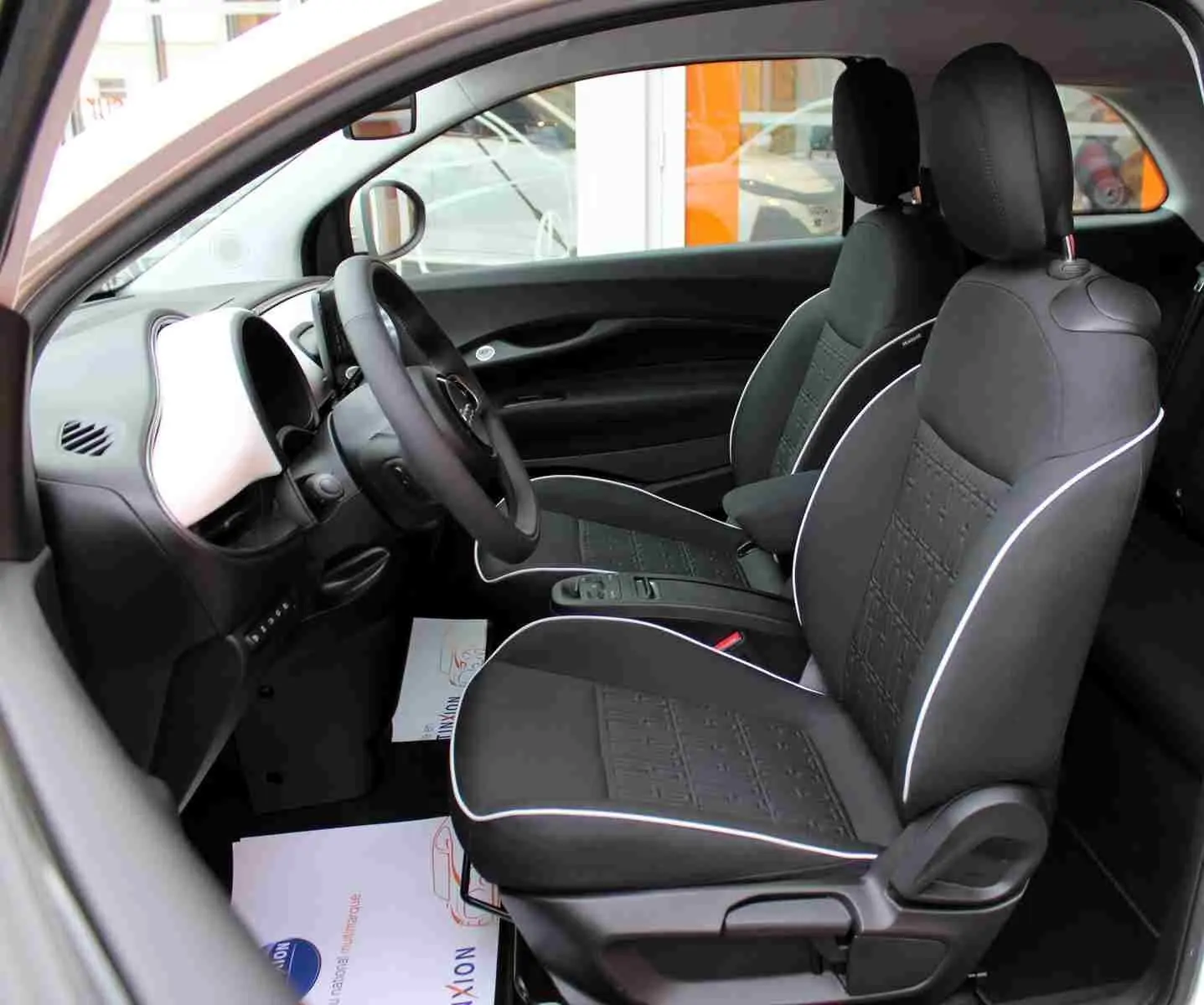 Vue intérieure côté gauche de la Fiat 500 électrique 2023, sièges noirs avec surpiqûres blanches et tableau de bord blanc.