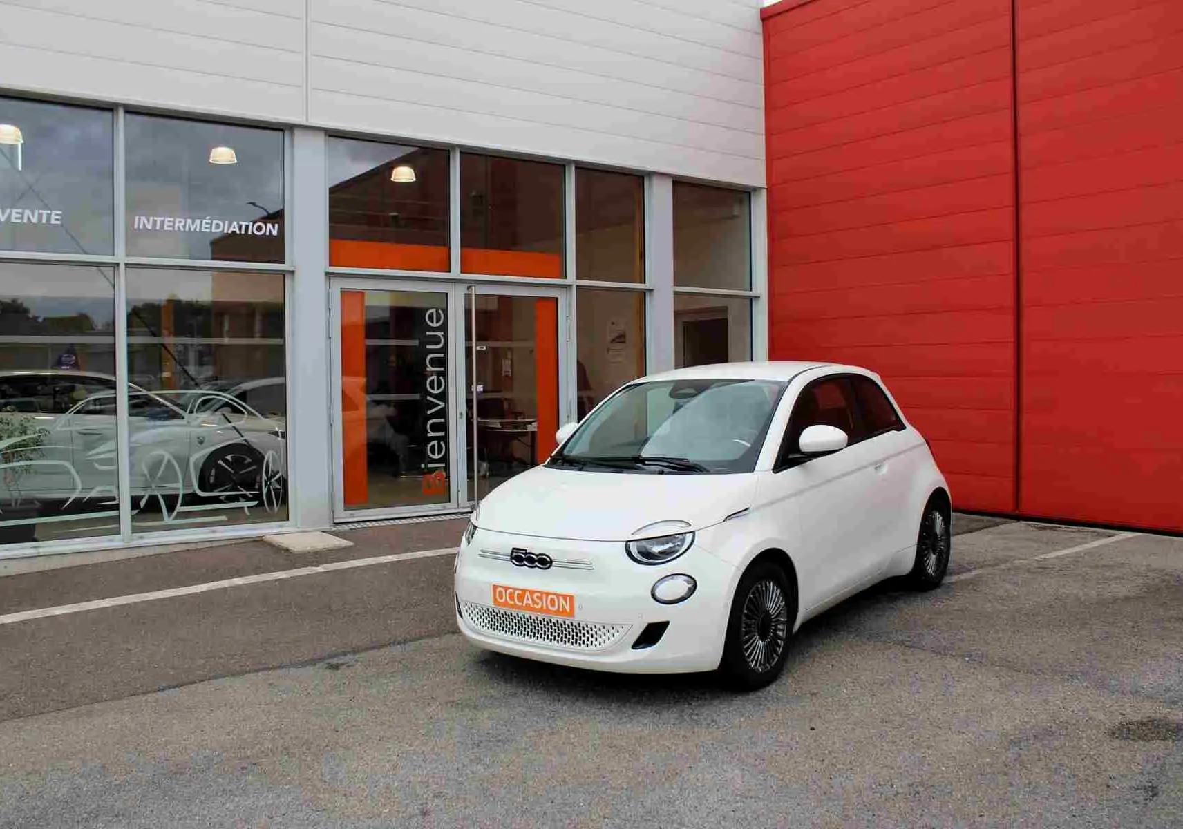 Fiat 500 électrique blanche vue 3/4 avant droit, stationnée devant un bâtiment avec panneau orange "Bienvenue".