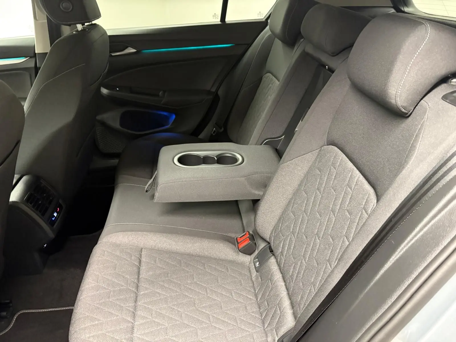 Vue intérieure arrière droite de la Volkswagen Golf 1.5 TSI 2025, sièges gris avec accoudoir central et éclairage d'ambiance bleu.