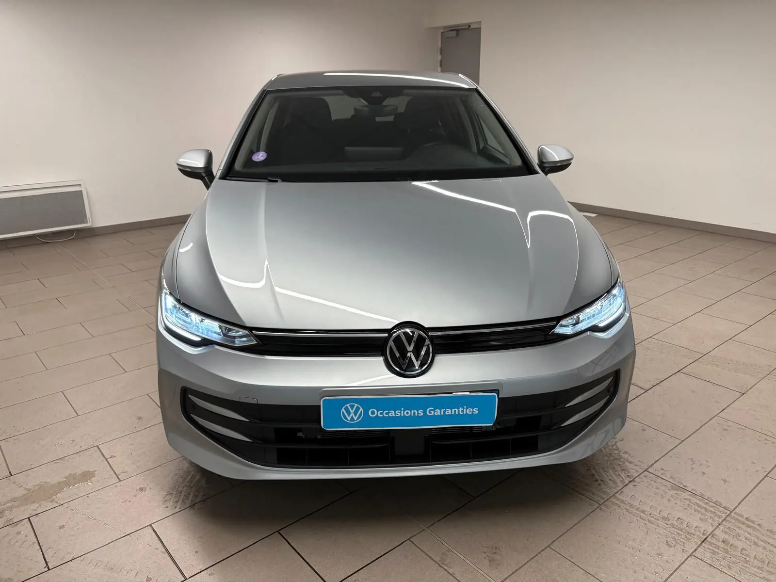 Vue avant d'une Volkswagen Golf 1.5 TSI 116 BVM6 VW Edition argent Dolomite avec phares LED allumés en intérieur.