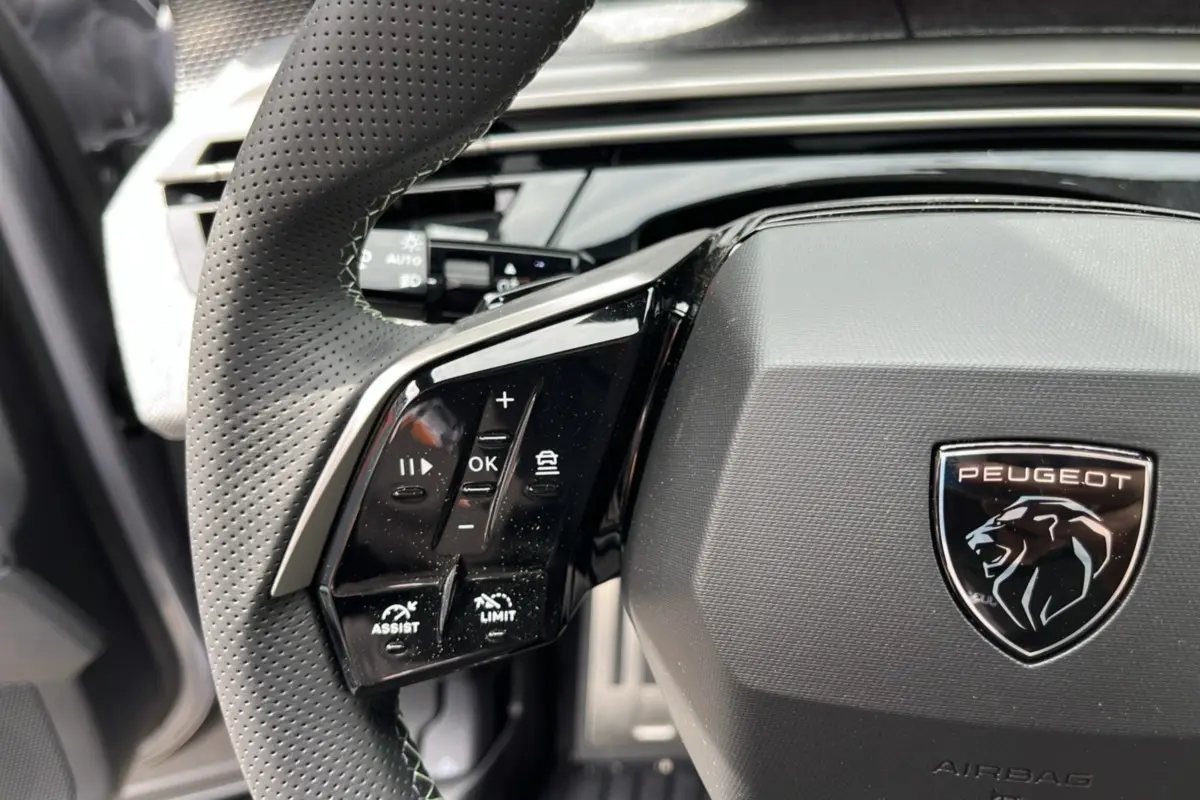Gros plan sur le volant cuir perforé avec commandes multifonctions et logo Peugeot sur fond gris du Peugeot 5008 Hybrid 2025.