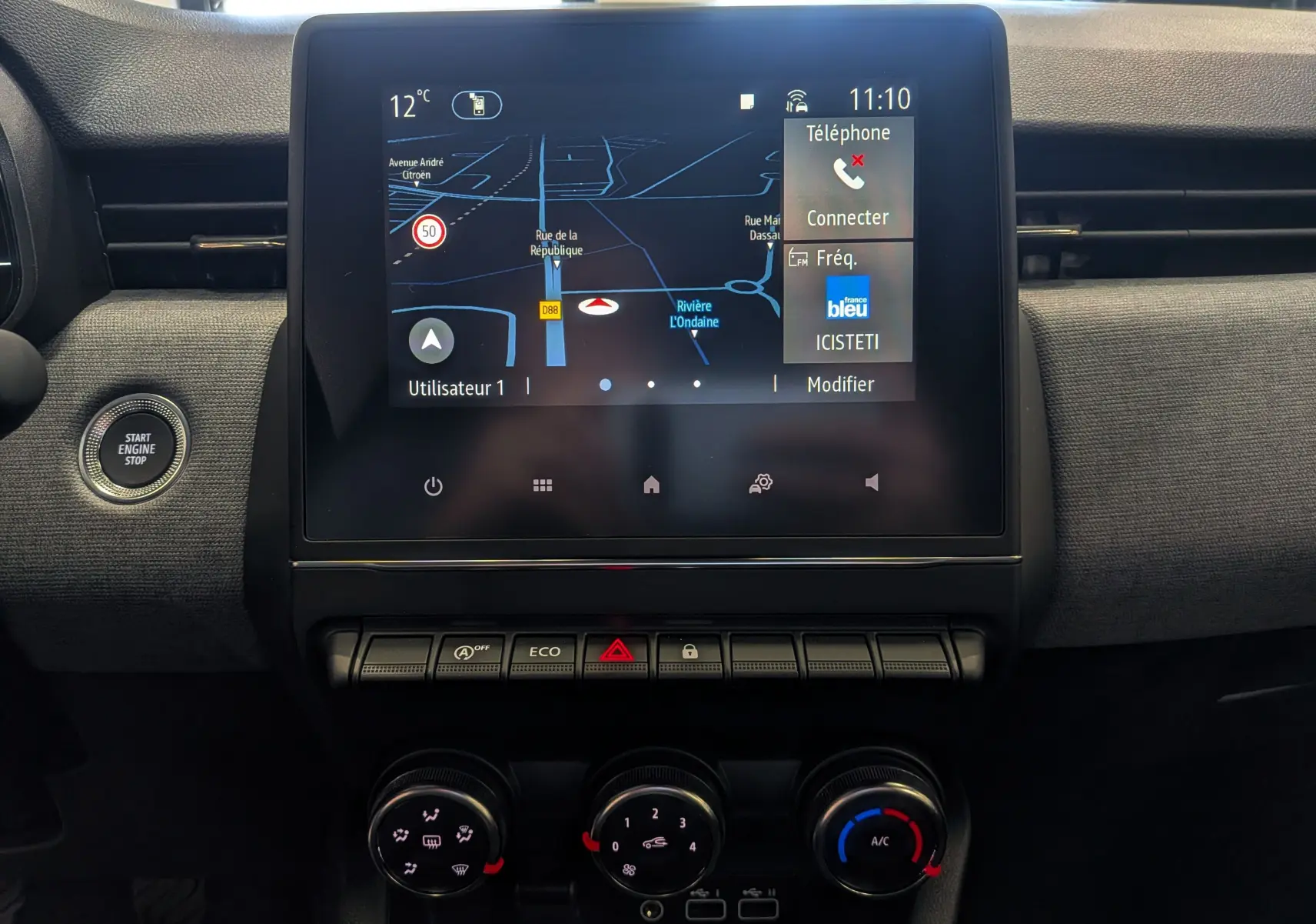 Vue intérieure centrée sur l’écran tactile du tableau de bord de la Renault Clio V gris Rafale avec commandes clim et bouton start visible.