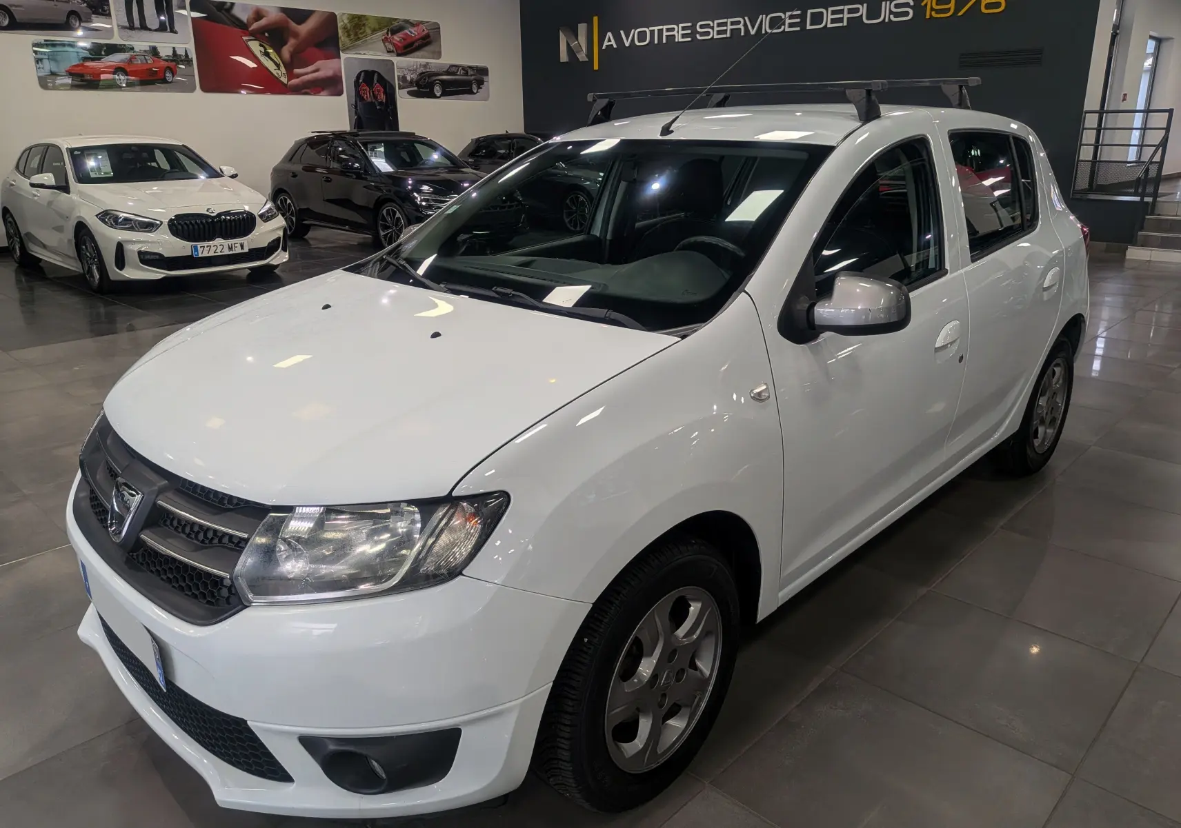 Vue 3/4 avant droite d'une Dacia Sandero TCe 90 blanche Glacier en showroom avec barres de toit noires.