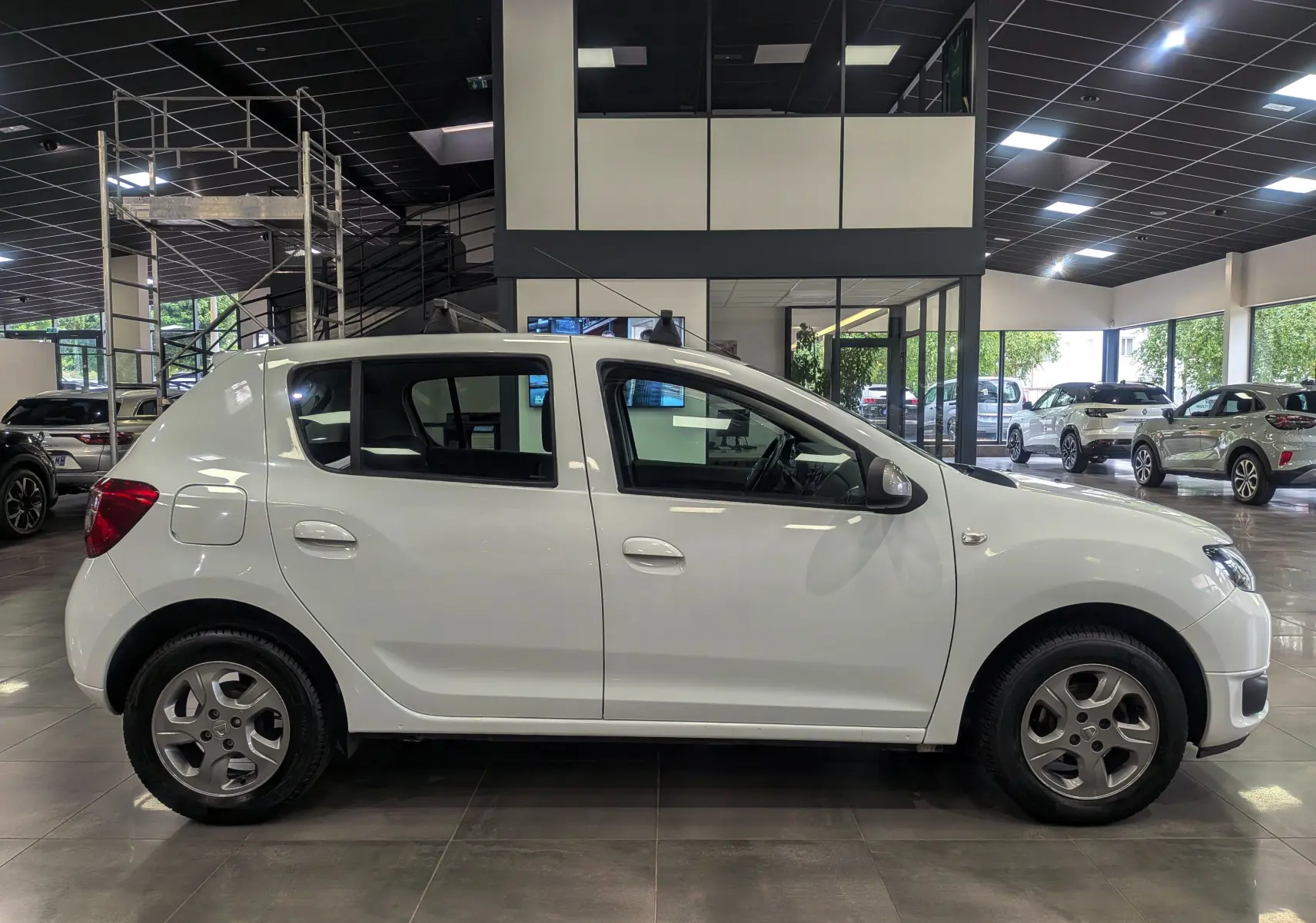 Profil côté gauche d'une Dacia Sandero blanc glacier de 2015, compacte et sobre, en intérieur de showroom.