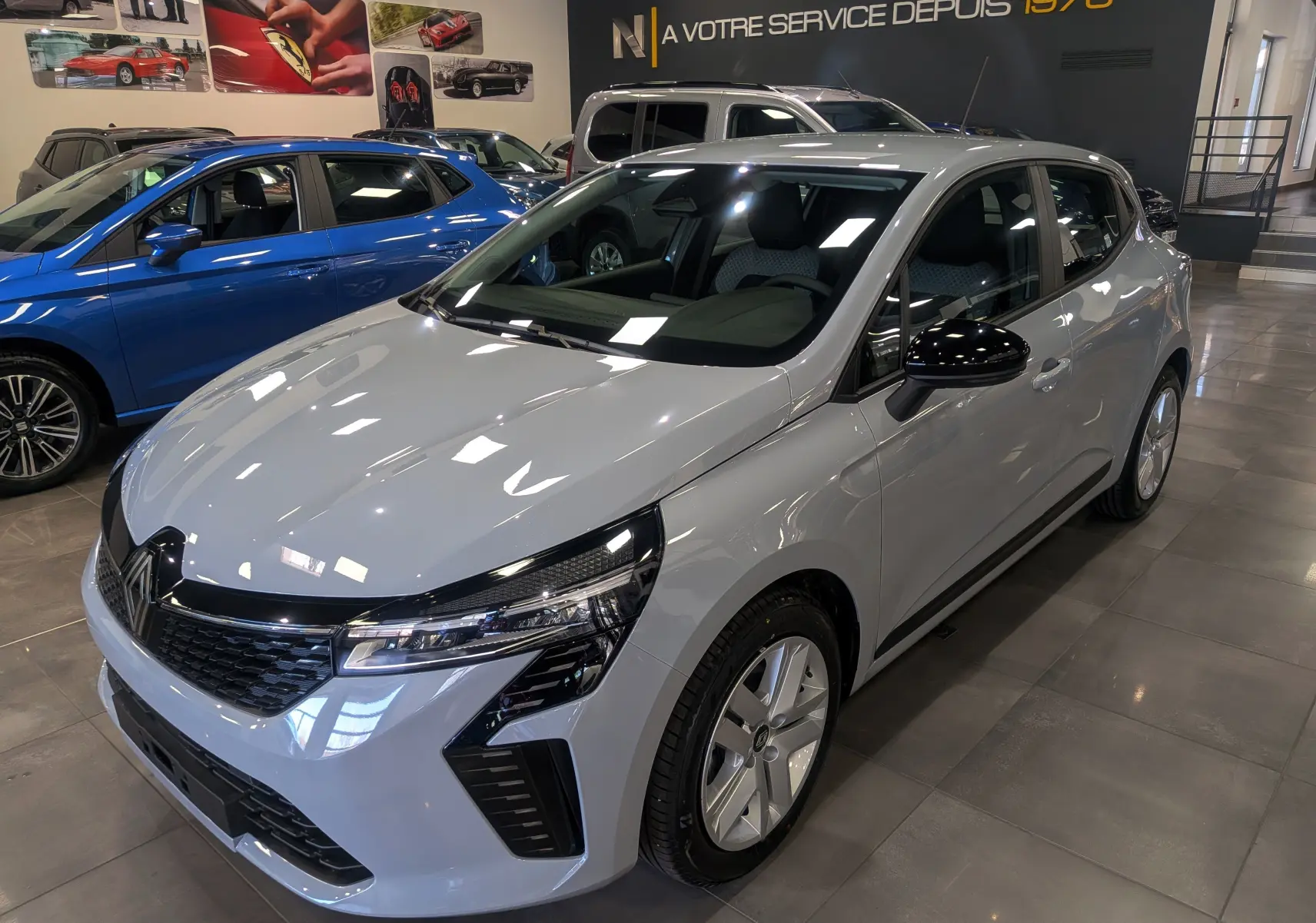 Vue 3/4 avant droite d'une Renault Clio V 2025 gris Rafale en showroom, avec rétroviseurs noirs et jantes spécifiques.