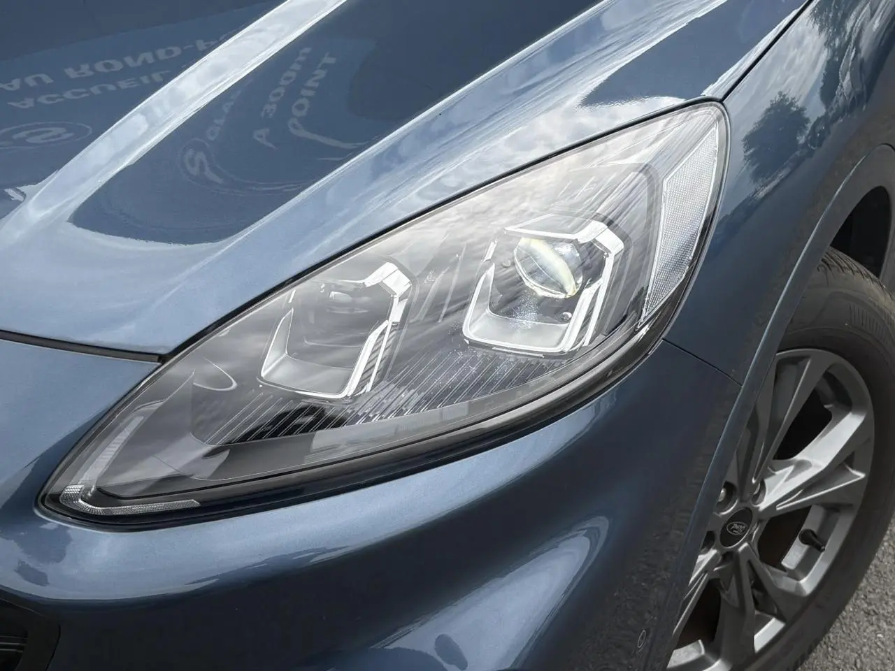 Gros plan sur le phare avant droit et la jante alliage 18 pouces du Ford Kuga Bleu Azur 2023 finition ST-Line.