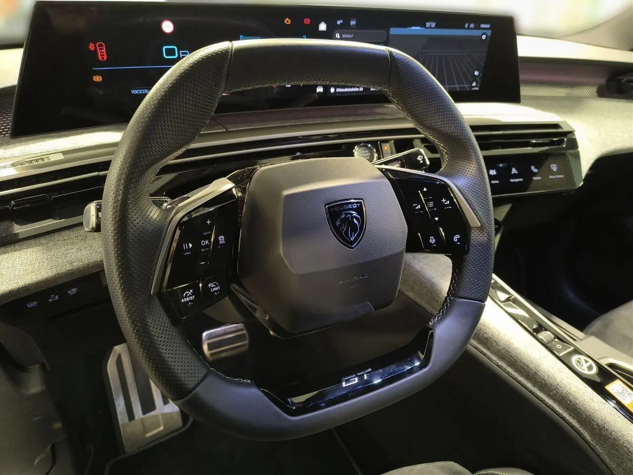 Vue rapprochée du volant compact noir perforé avec badge Peugeot et inscription GT, tableau de bord numérique en arrière-plan.