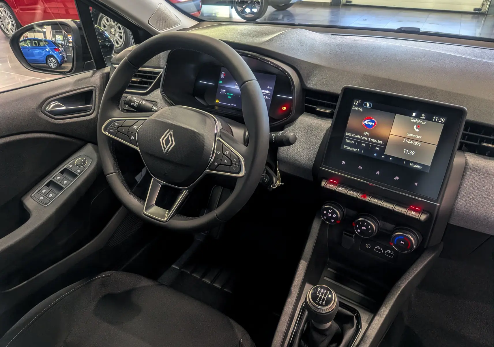 Vue intérieure côté conducteur de la Renault Clio V 2025, tableau de bord moderne avec écran tactile et levier de vitesse manuel.