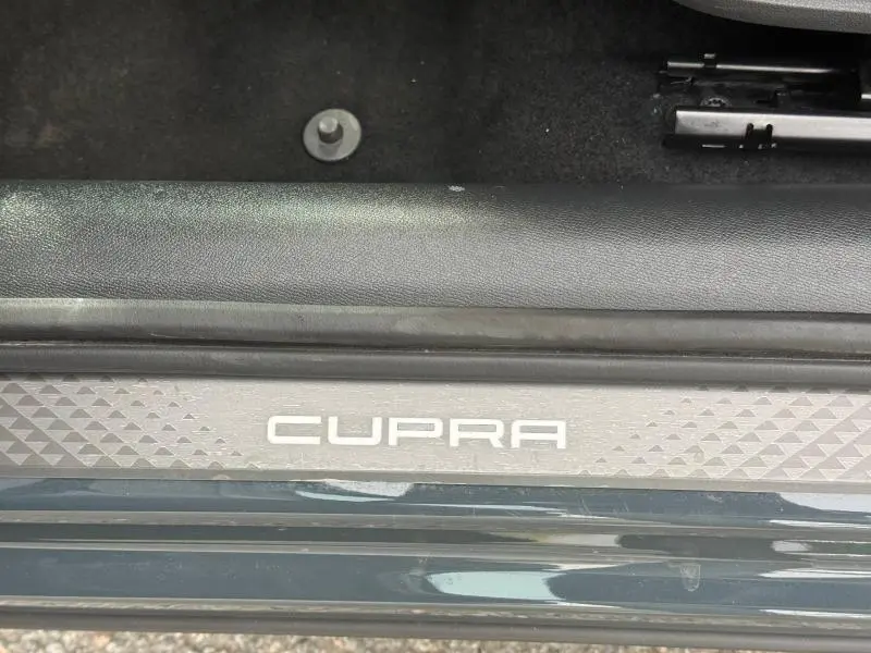 Gros plan sur la plaque de seuil de porte noire avec logo CUPRA blanc, intérieur moquette noire visible.