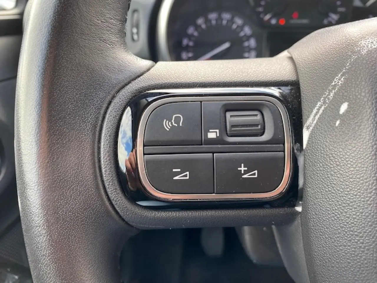 Gros plan sur les commandes de volume et téléphone au volant d'une Citroën C3 gris clair, intérieur visible en arrière-plan flou.