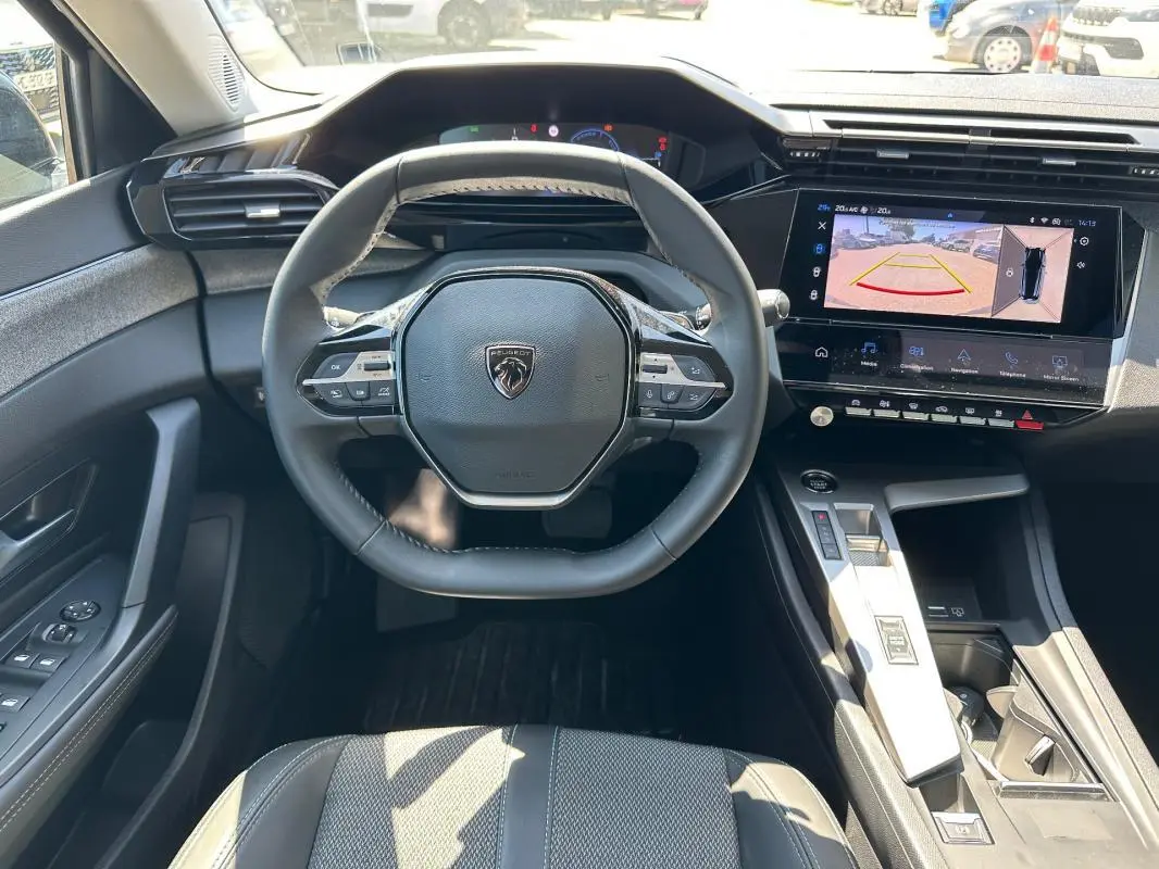 Vue intérieure centrée sur le volant et la console d'une Peugeot 308 SW Hybrid noire avec écran tactile et caméra de recul affichée.
