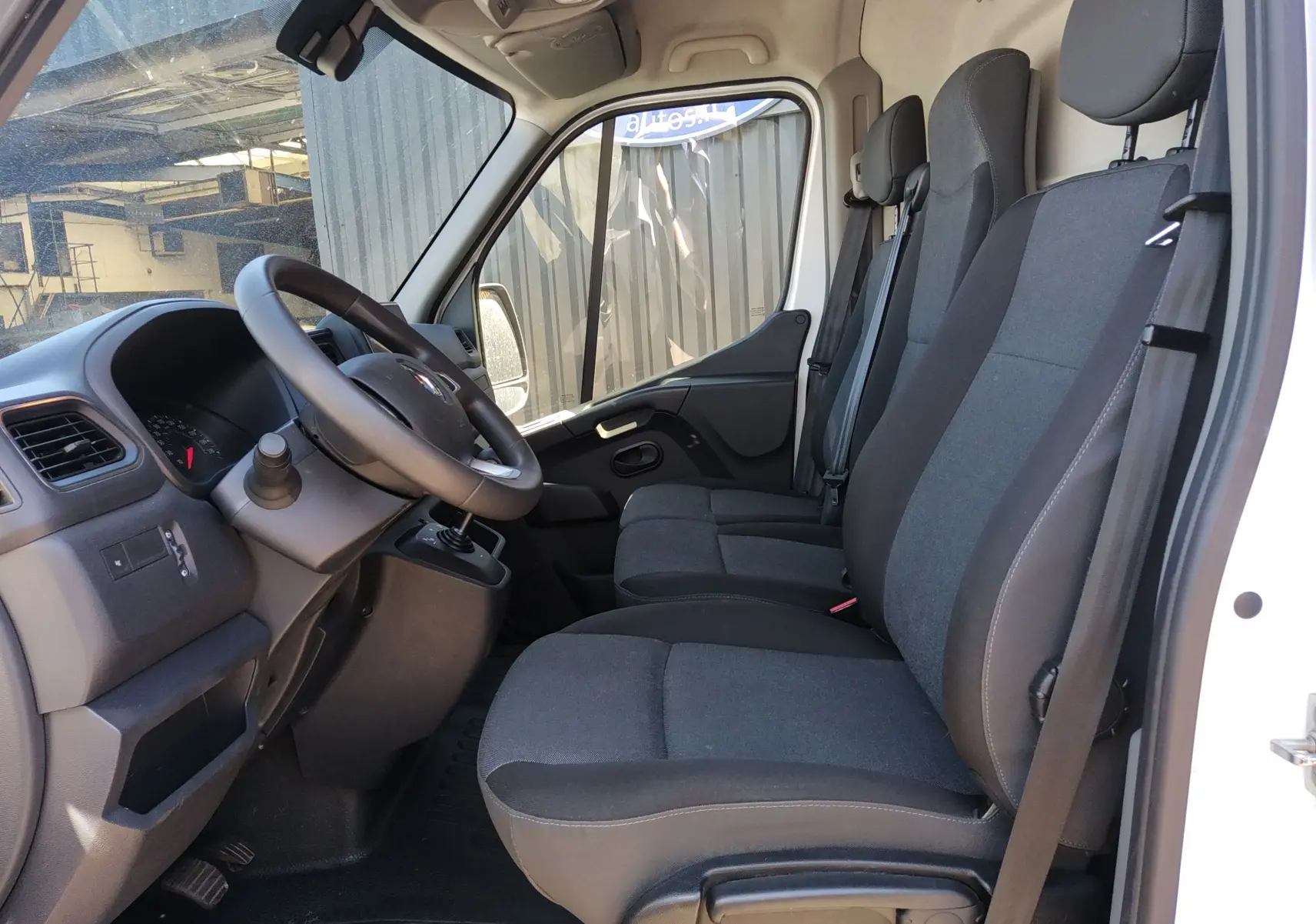 Intérieur du Renault Master III blanc, vue côté gauche montrant le poste de conduite et les trois sièges en tissu noir.