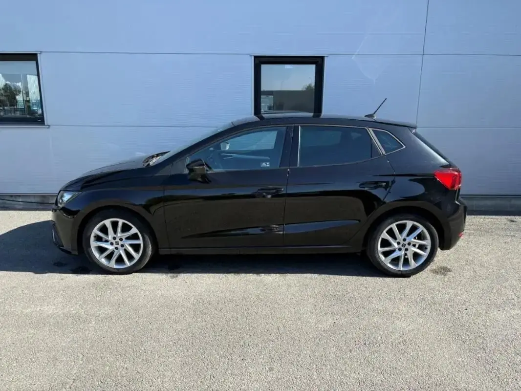 Vue latérale droite d'une SEAT Ibiza 1.0 TSI 95 FR noire, avec jantes alliage et vitres teintées, stationnée devant un mur gris.