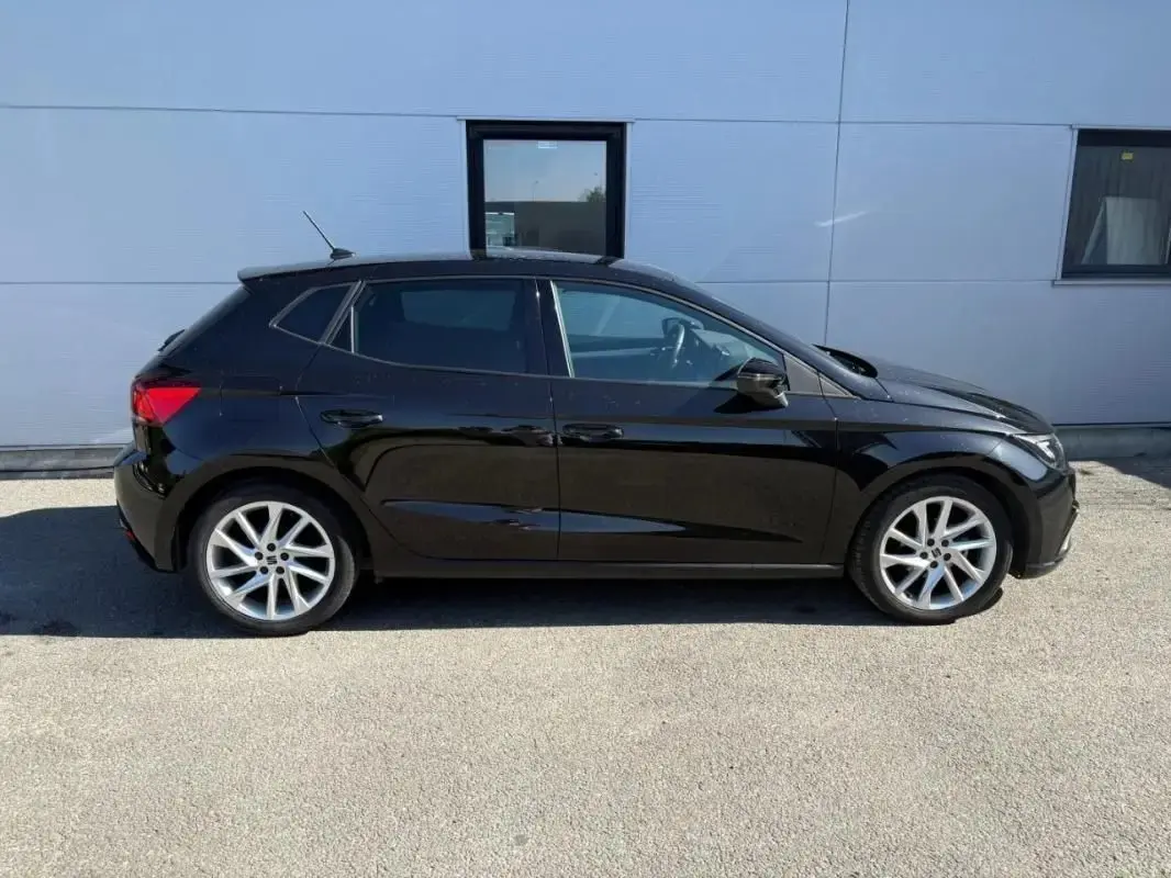 Profil droit d’une SEAT Ibiza 1.0 TSI 95 FR noire, avec jantes alliage argentées, stationnée devant un mur gris.