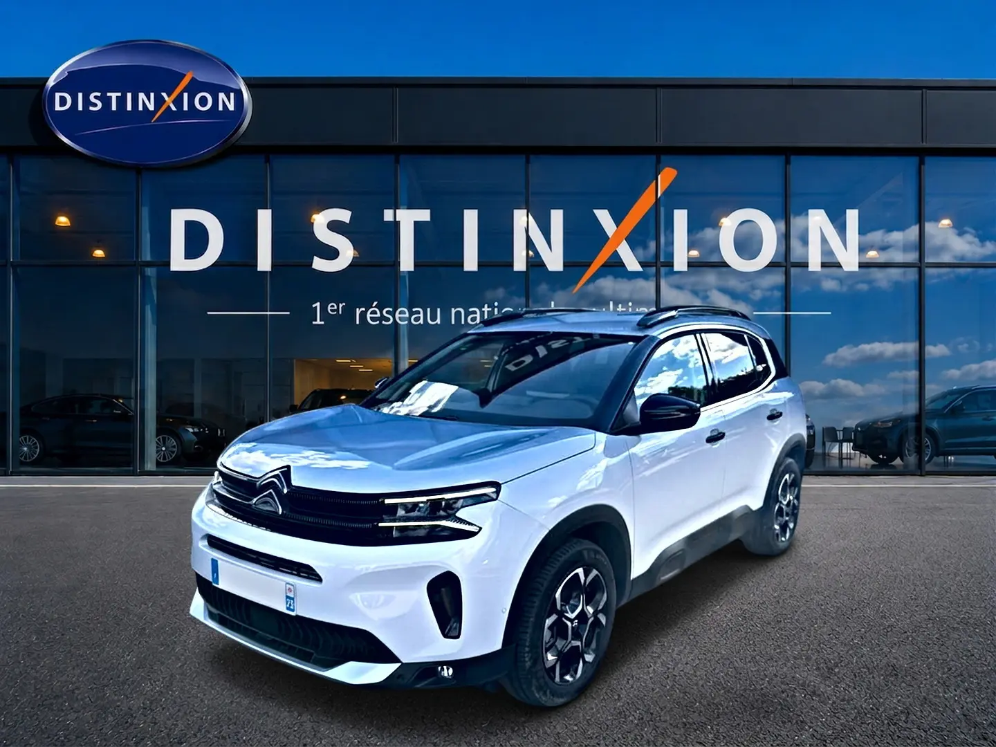 Citroën C5 Aircross blanc en 3/4 avant droit, avec jantes alu et barres de toit visibles devant un showroom.