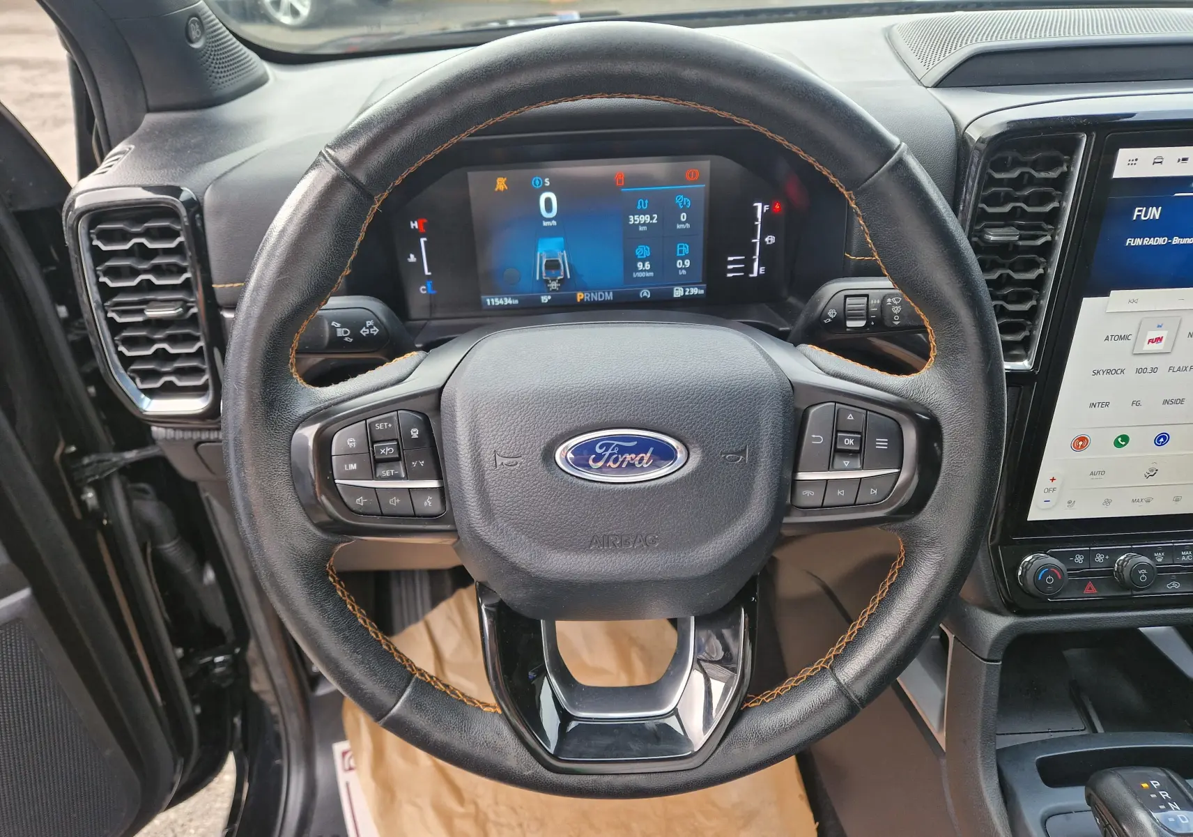 Vue intérieure centrée sur le volant noir en cuir avec surpiqûres orange du Ford Ranger Wildtrak 2023, tableau de bord digital visible.