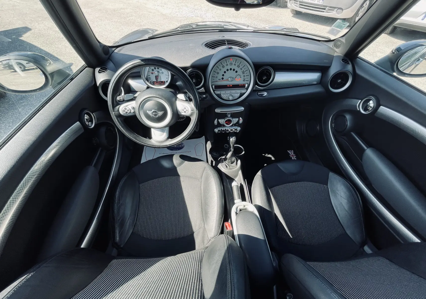 Vue intérieure avant de la MINI Cooper S 2008 avec tableau de bord rond, volant multifonction et sièges noirs en tissu et cuir.