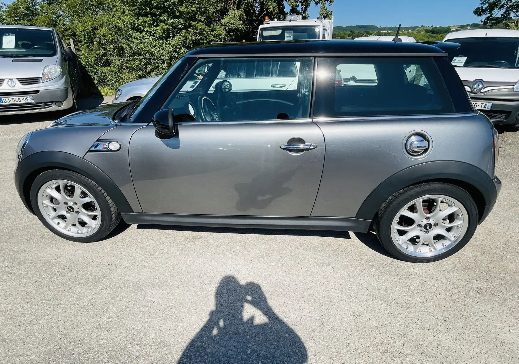 Profil côté gauche d'une MINI Cooper S gris métallisé avec toit noir et jantes alliage sur un parking en extérieur.