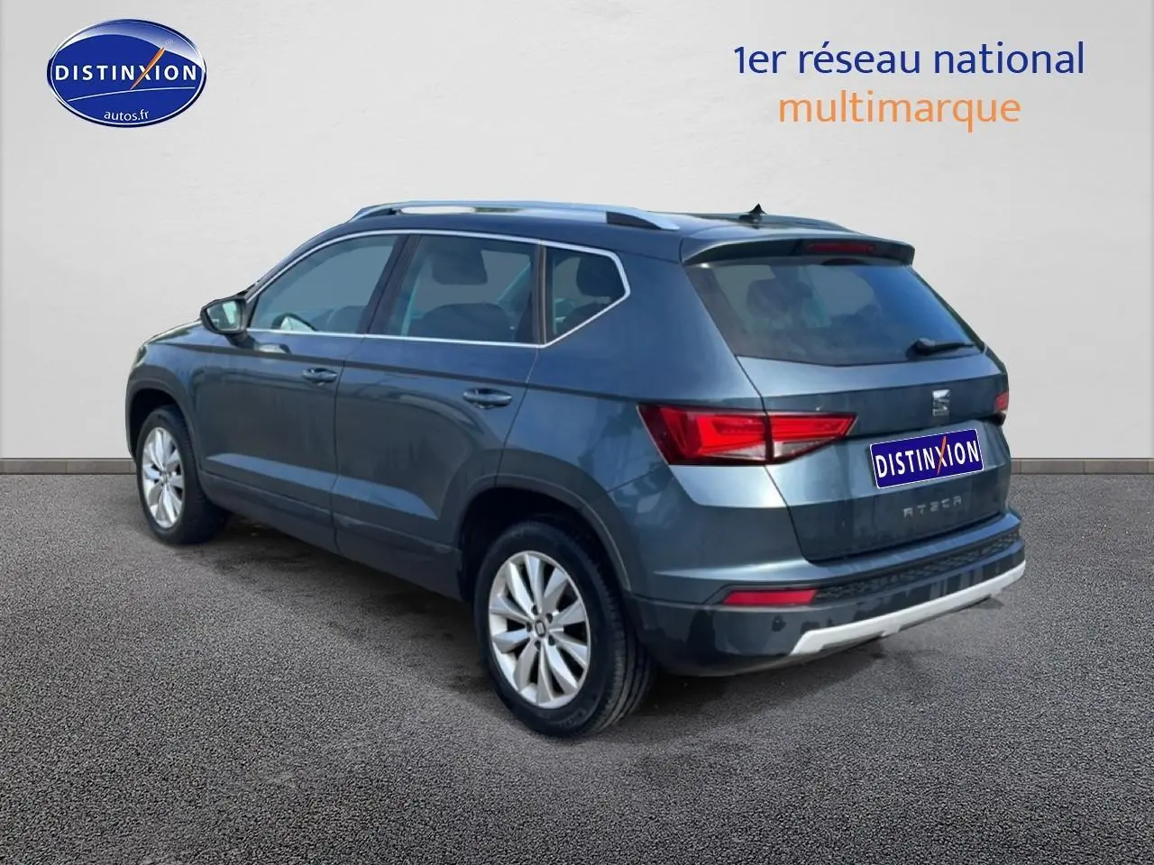 Vue 3/4 arrière droite d'un SEAT Ateca bleu/gris avec jantes alu et barres de toit visibles sur fond neutre.