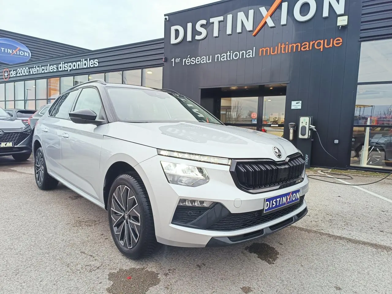 Vue 3/4 avant d’un Skoda Kamiq 2025 argent avec toit noir et jantes alliage 17 pouces, devant un showroom Distinxion.