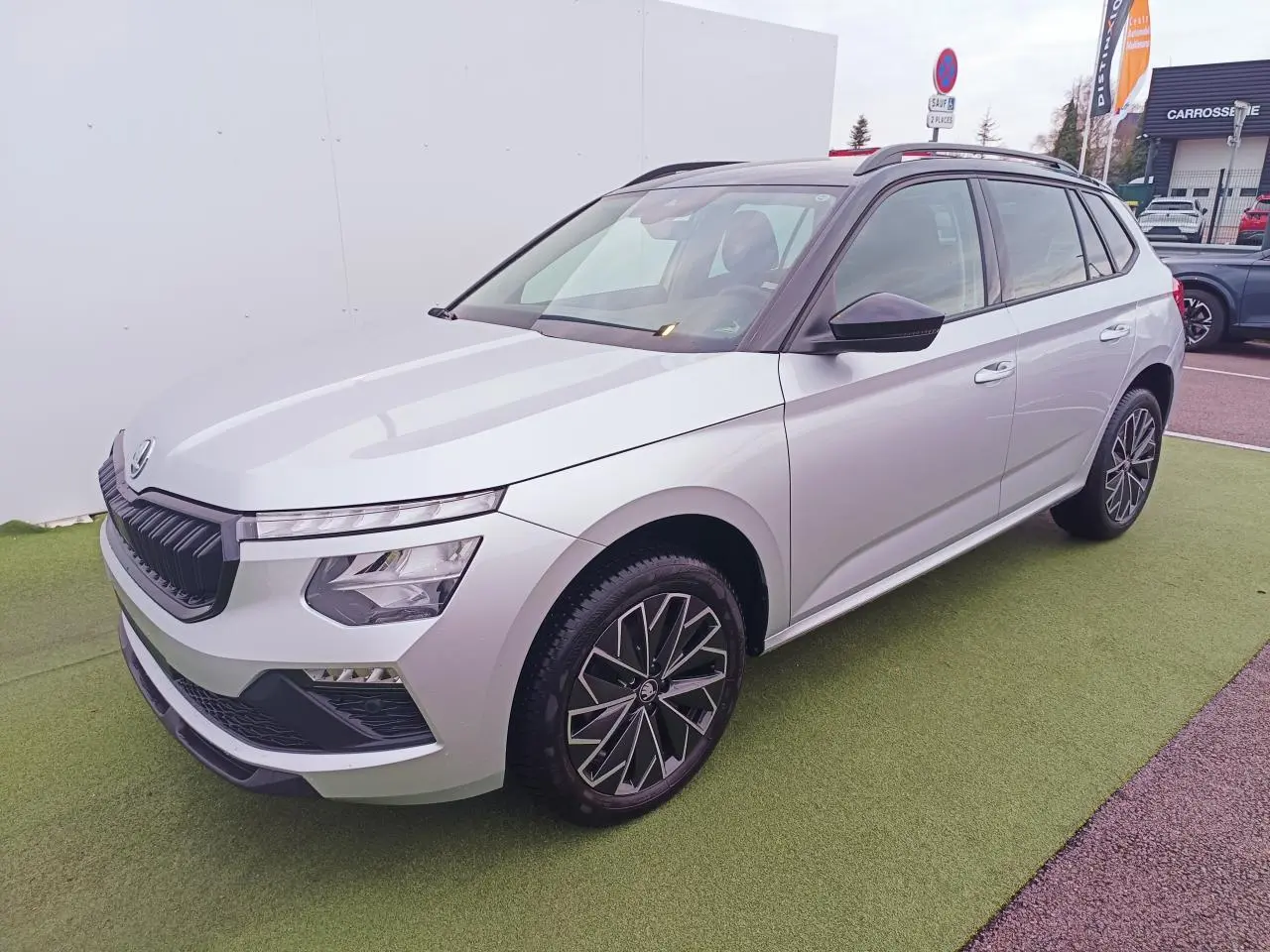 Vue 3/4 avant d'un Skoda Kamiq 2025 argent avec toit noir et jantes alliage 17 pouces, stationné sur sol vert.