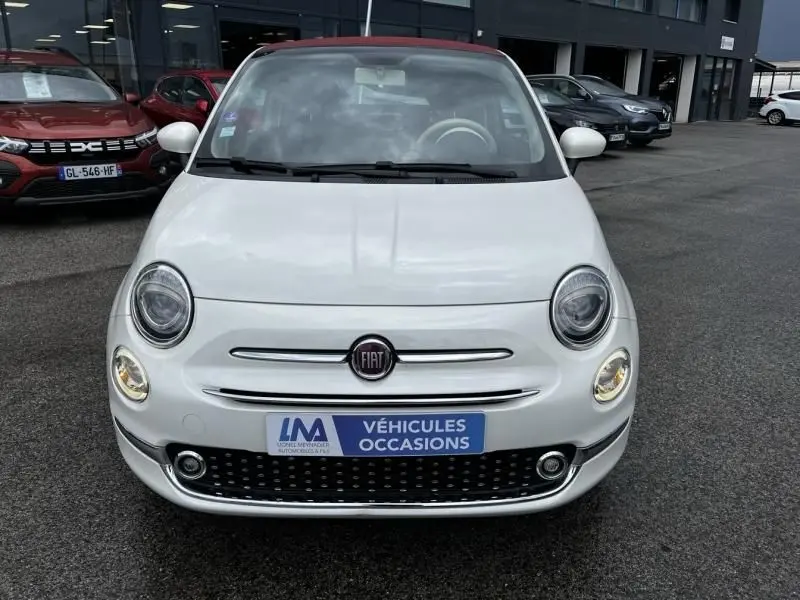 Vue frontale d'une FIAT 500C blanche 2020 avec toit rouge et phares ronds allumés sur parking de véhicules d'occasion.