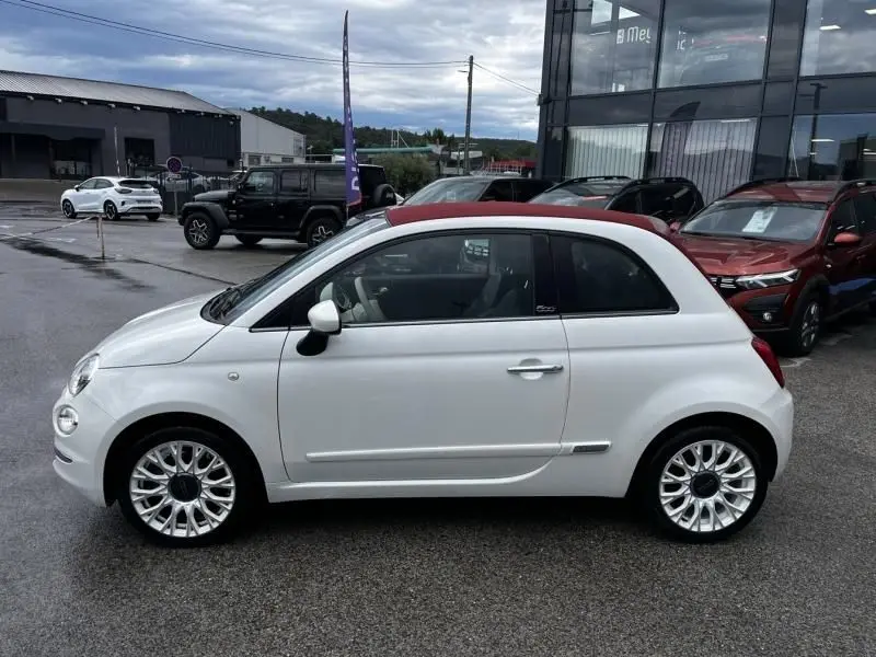 Vue latérale droite d'une Fiat 500C blanche 2020 avec toit ouvrant rouge, garée devant un bâtiment vitré.