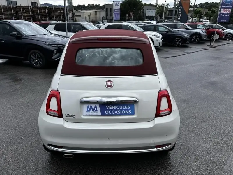 Vue arrière d'une FIAT 500C blanche avec toit ouvrant rouge bordeaux sur un parking de véhicules d'occasion.