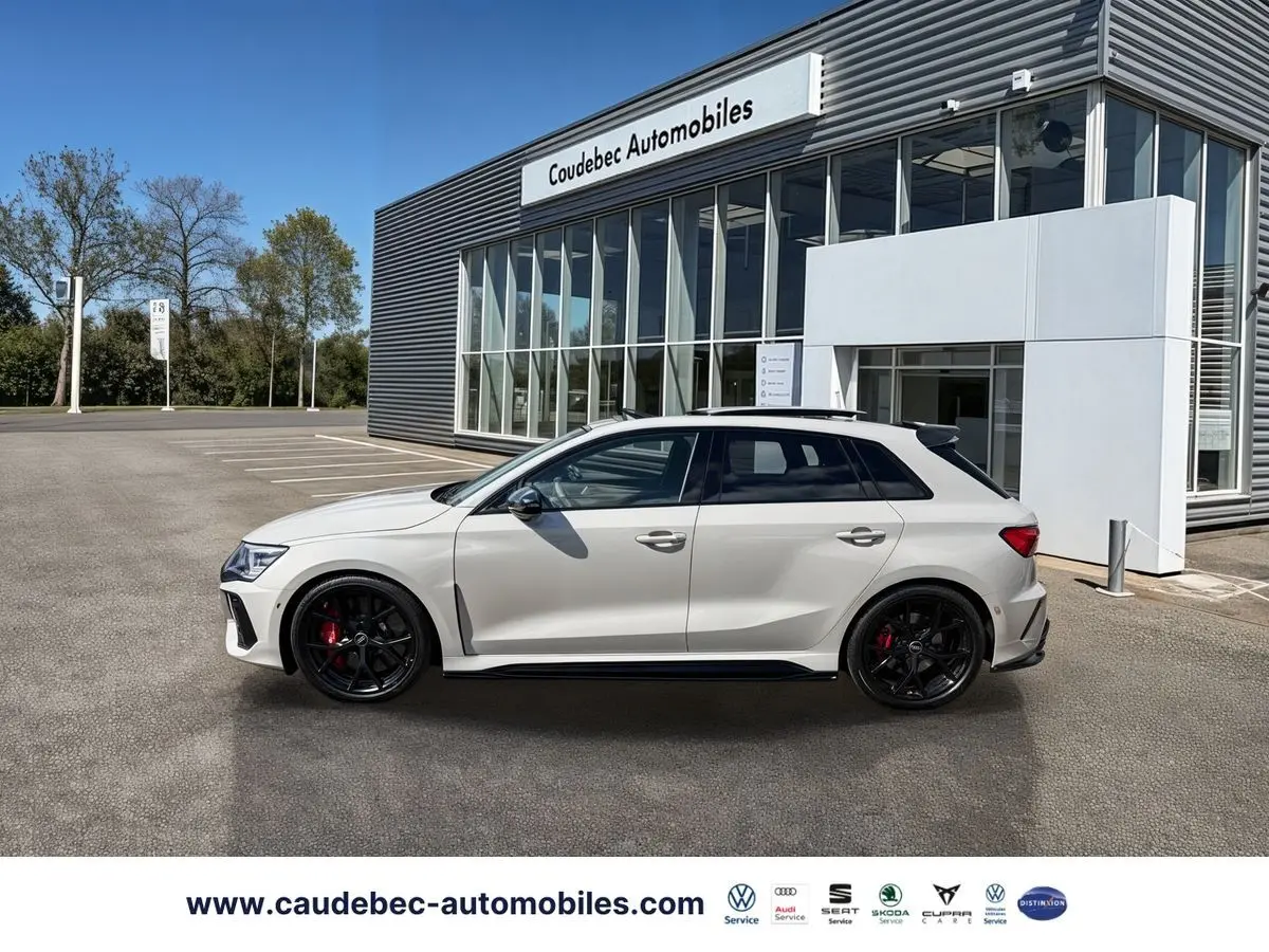 Audi RS3 Sportback gris vue de profil droit, jantes noires et étriers de frein rouges, devant un bâtiment moderne.
