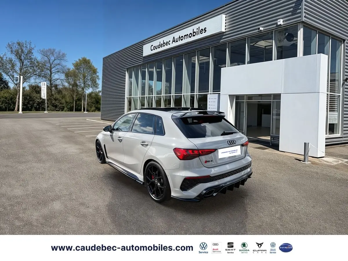 Audi RS3 Sportback gris vue 3/4 arrière droit, jantes noires, toit panoramique et becquet carbone visible.