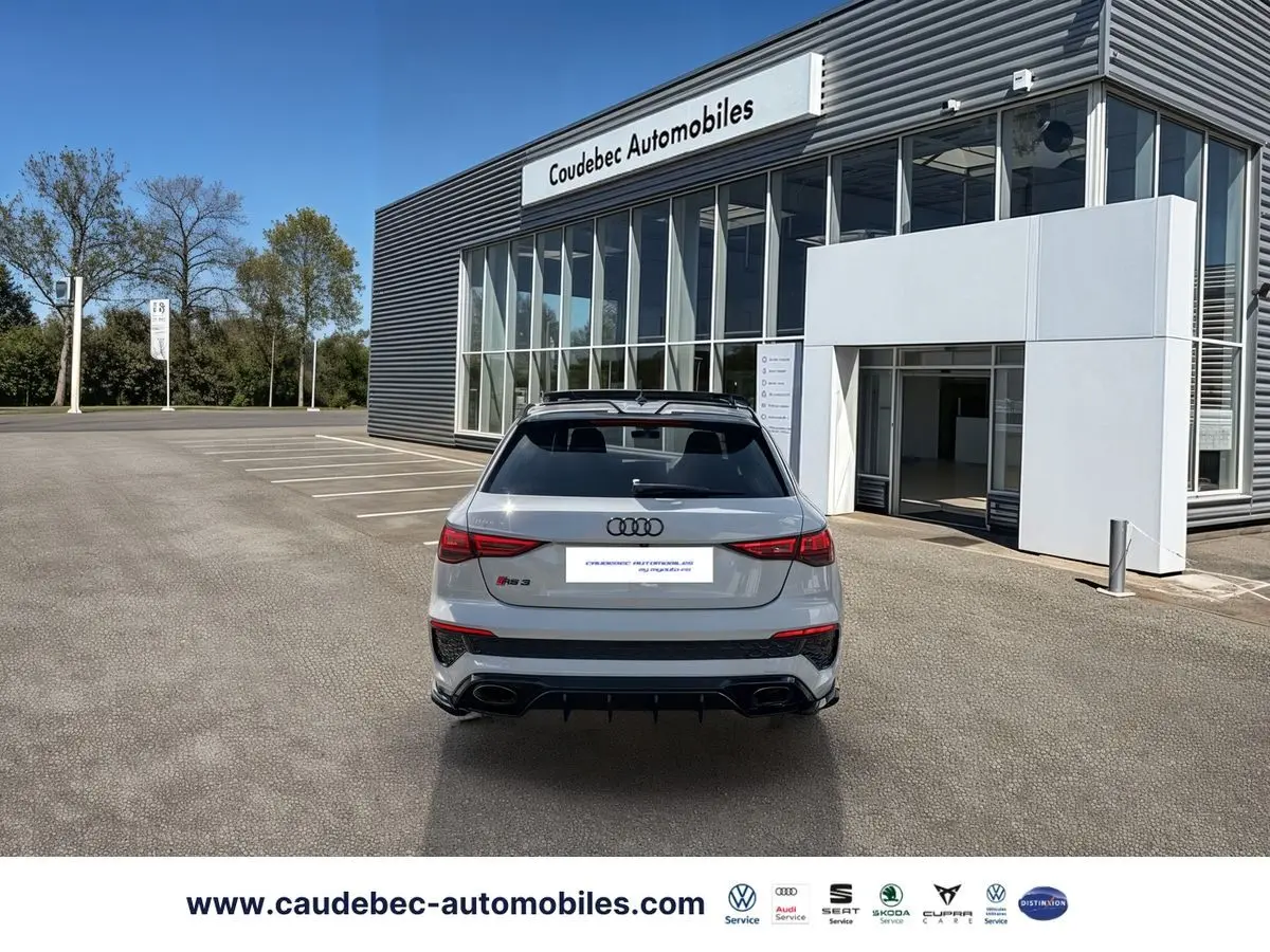 Vue arrière d'une Audi RS3 Sportback grise avec diffuseur noir et feux arrière LED, stationnée devant un bâtiment moderne.