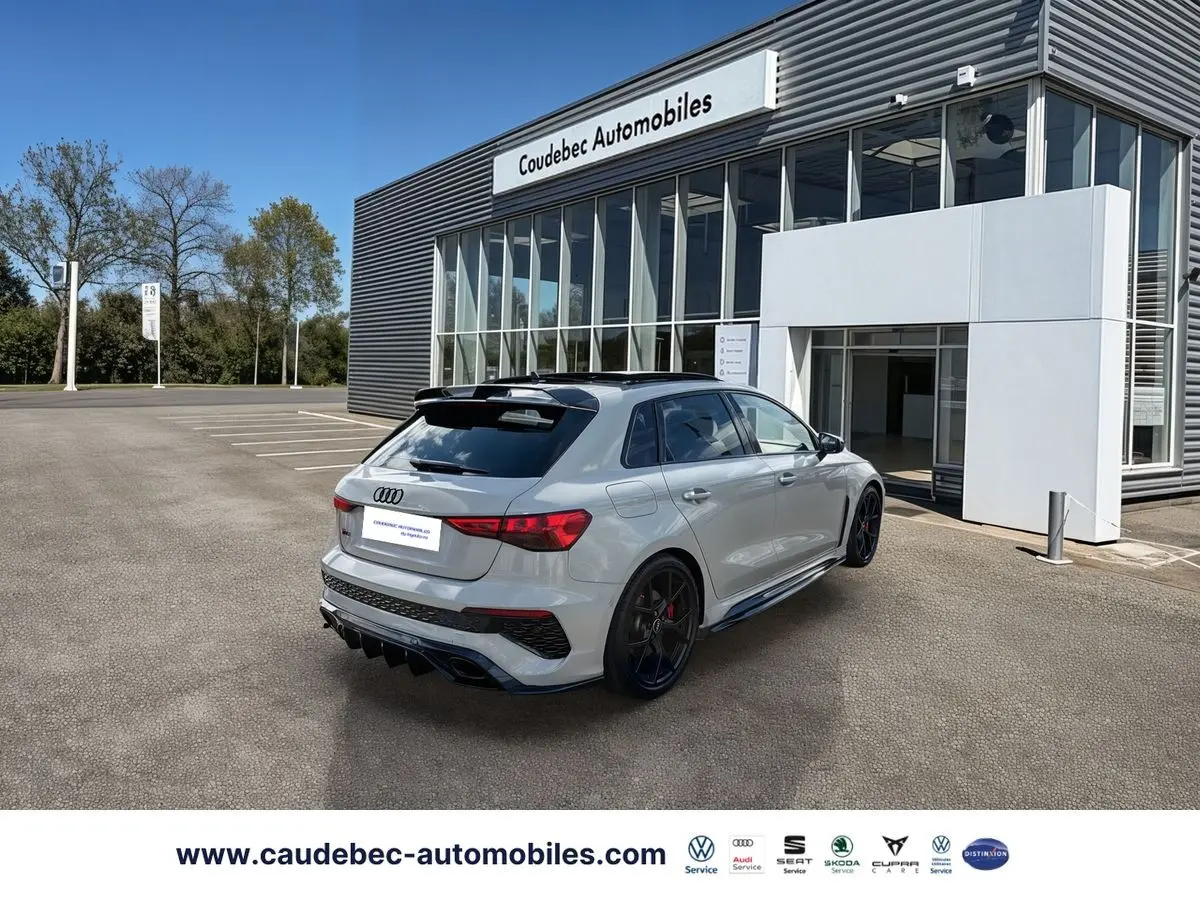 Audi RS3 Sportback grise vue 3/4 arrière droit, jantes noires mat et détails carbone visibles, stationnée devant un showroom.