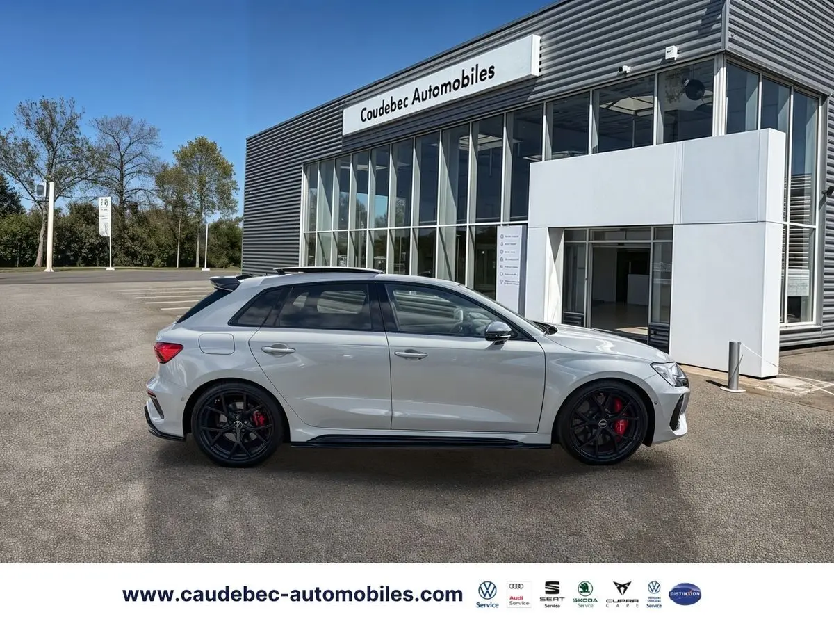 Audi RS3 Sportback grise vue de profil côté gauche, avec jantes noires et étriers de frein rouges, devant un showroom.