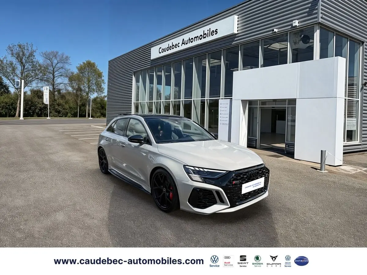 Audi RS3 Sportback gris vue 3/4 avant droit, jantes noires et étriers de frein rouges devant un showroom moderne.