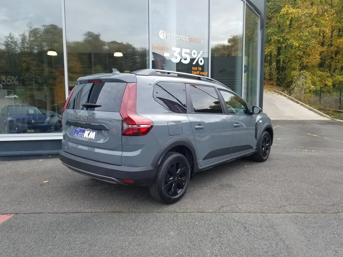 Vue 3/4 arrière droite d'un Dacia Jogger gris urbain avec barres de toit et vitres teintées, stationné devant une vitrine.