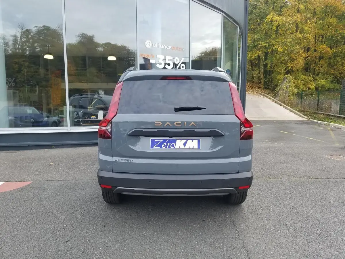 Vue arrière d'un Dacia Jogger gris urbain 2025 avec feux verticaux et logo doré sur un parking devant une vitrine.