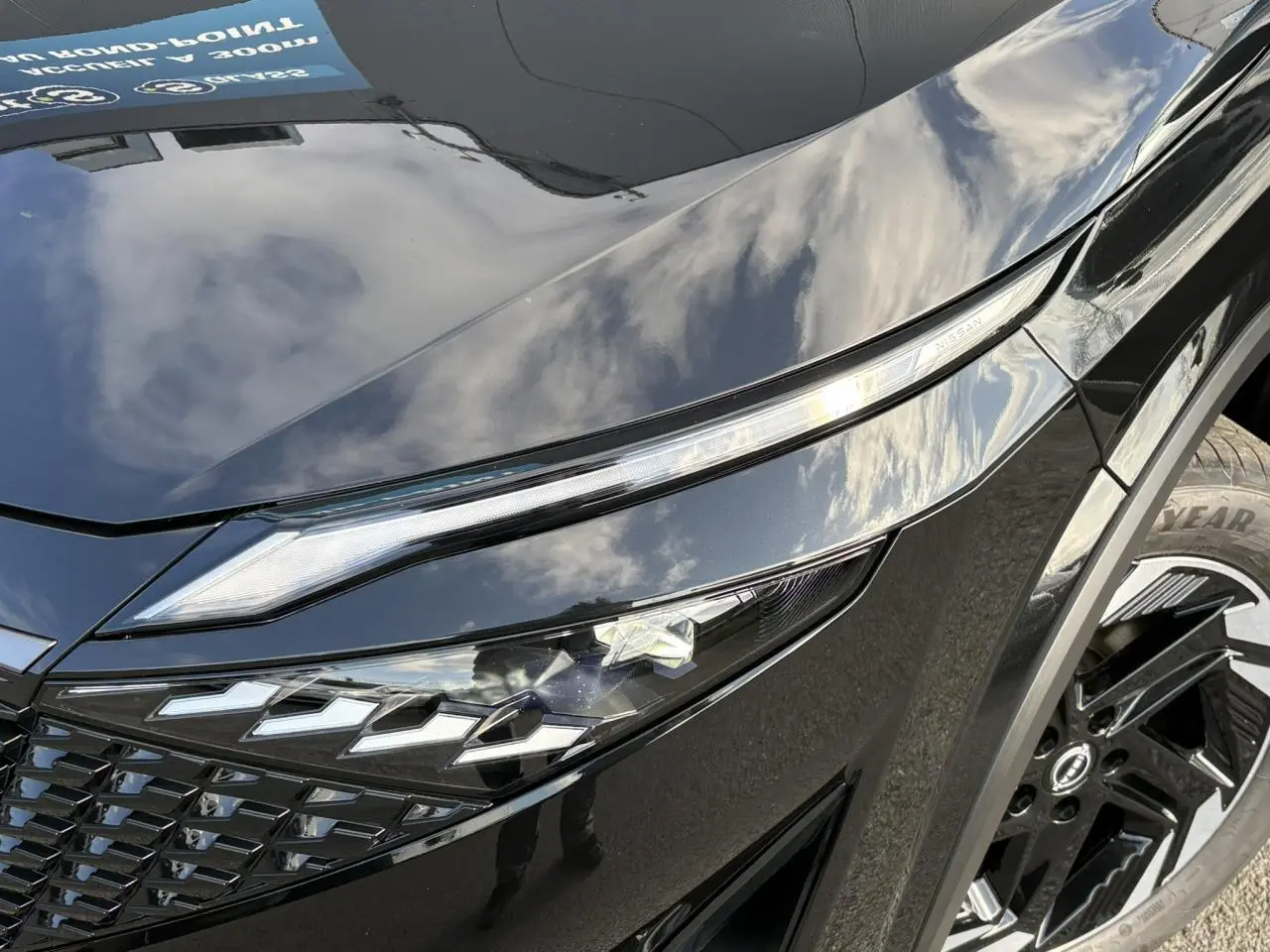 Gros plan sur le phare avant gauche et la jante noire du Nissan Qashqai III e-Power 2025 en peinture métallisée noire.