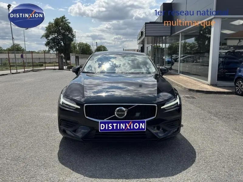 Vue avant d'une Volvo S60 2024 noire brillante avec calandre distinctive et phares LED allumés, stationnée devant un showroom.