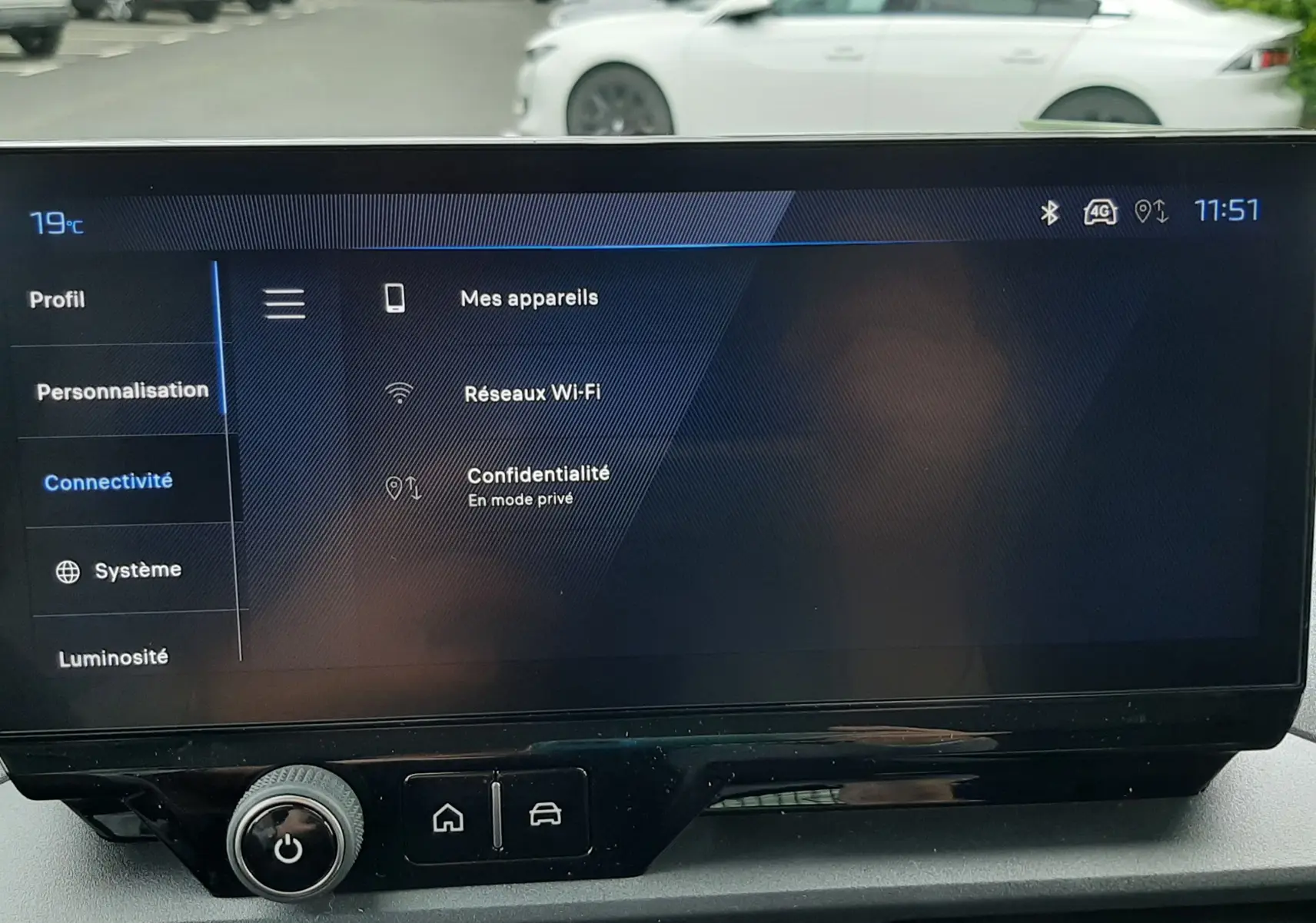 Vue intérieure centrée sur l'écran tactile du tableau de bord du Peugeot Rifter, avec vue floue d'une voiture blanche en arrière-plan.